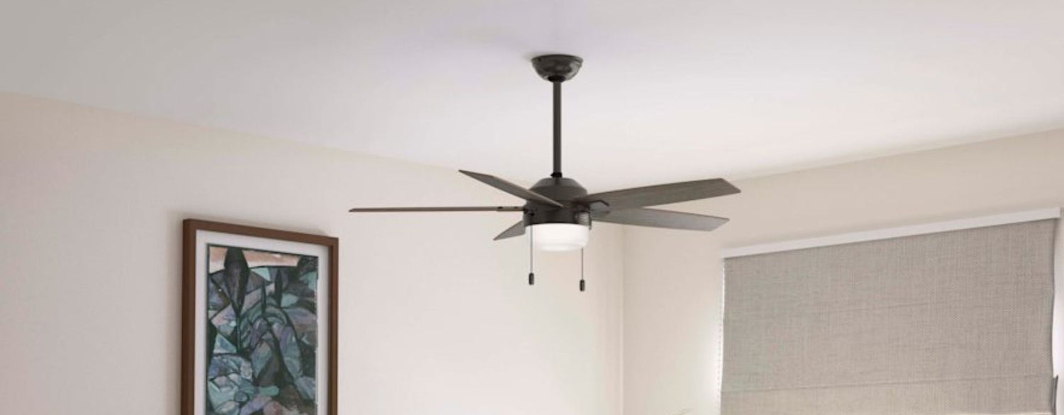 Lifestyle image of a contemporary bedroom featuring a dark bronze/black ceiling fan with five blades, an integrated light, and dual pull chains. The fan is installed on a downrod above a light, airy bedroom with warm wood furniture, neutral bedding, and indoor greenery, showcasing a blend of modern functionality and transitional style.