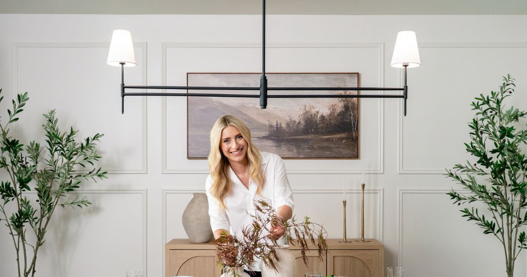 Dining room lifestyle image featuring a woman styling a table beneath a modern black linear light fixture with two white shades, framed by warm neutral décor and greenery.