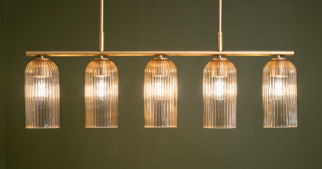 Five-light linear chandelier with ribbed glass shades and a warm gold finish against a dark green wall.