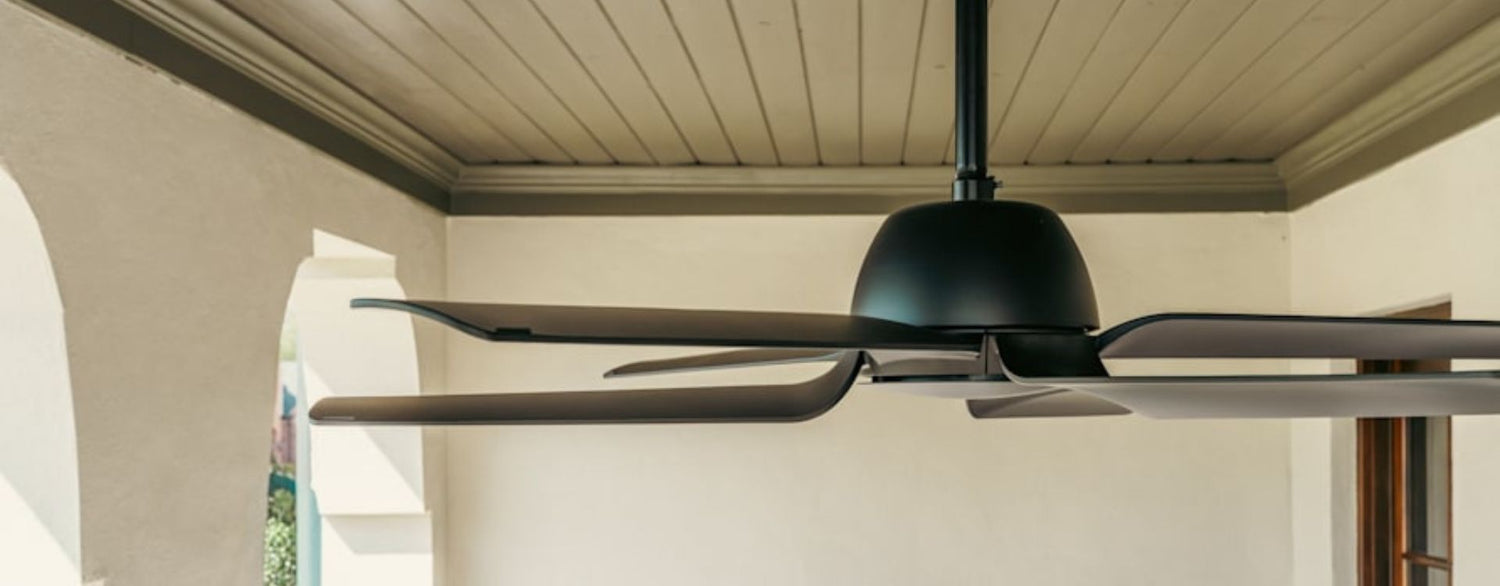Close-up lifestyle image of a contemporary outdoor ceiling fan showcasing its distinctive curved blade design, matte black finish, and architectural silhouette beneath a covered porch ceiling.