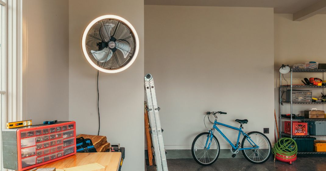 Garage lifestyle image featuring a wall-mounted Hunter garage fan with an illuminated outer ring, alongside a workbench, ladder, bicycle, and storage shelving.