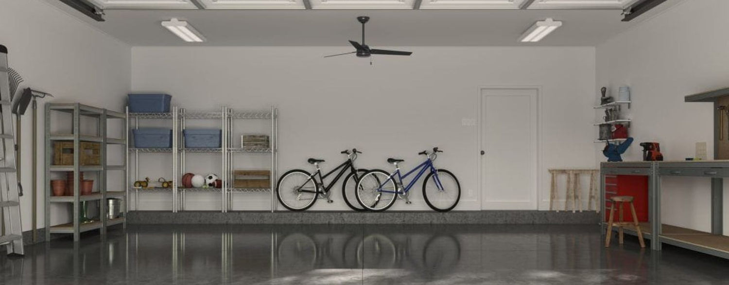 Wide lifestyle image of a clean, organized residential garage featuring a black ceiling fan centered overhead. Storage shelving, bicycles, a workbench, and utility lighting establish the garage setting while highlighting the fan as a practical solution for improving airflow and comfort.