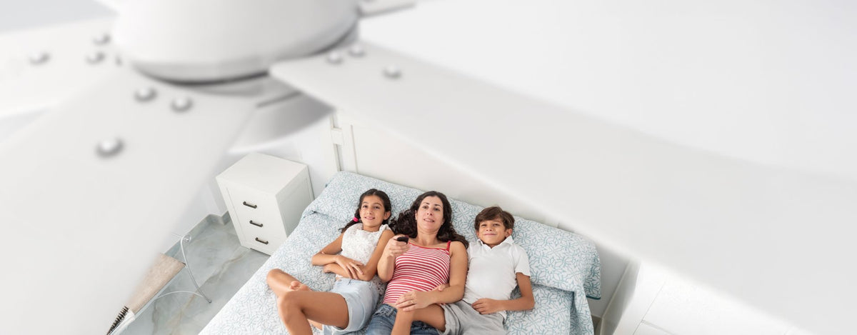 Overhead view of a mother and two children lying on a bed, comfortably enjoying airflow from a ceiling fan while using a remote.