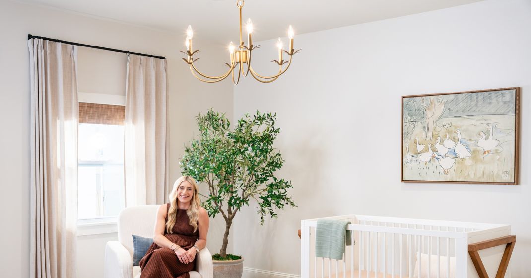 Bright nursery lifestyle image featuring a woman seated beside a crib, with a gold chandelier overhead and a large indoor tree adding warmth to the room.